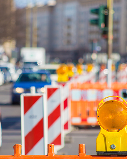 Fahrzeuge fahren an einer Straßenbaustelle in der Stadt vorbei. Ein Radfahrer (rechts) will die Straße überqueren. © Rainer Fuhrmann - stock.adobe.com