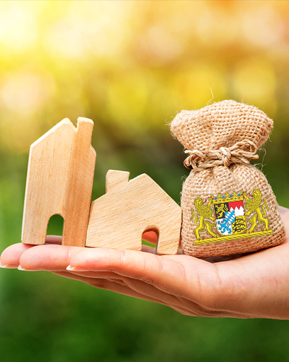 Hand hält Geldbeutel (links) und ein Musterhaus (rechts). Auf dem Geldbeutel ist das bayerische Staatswappen. © Watchara - stock.adobe.com