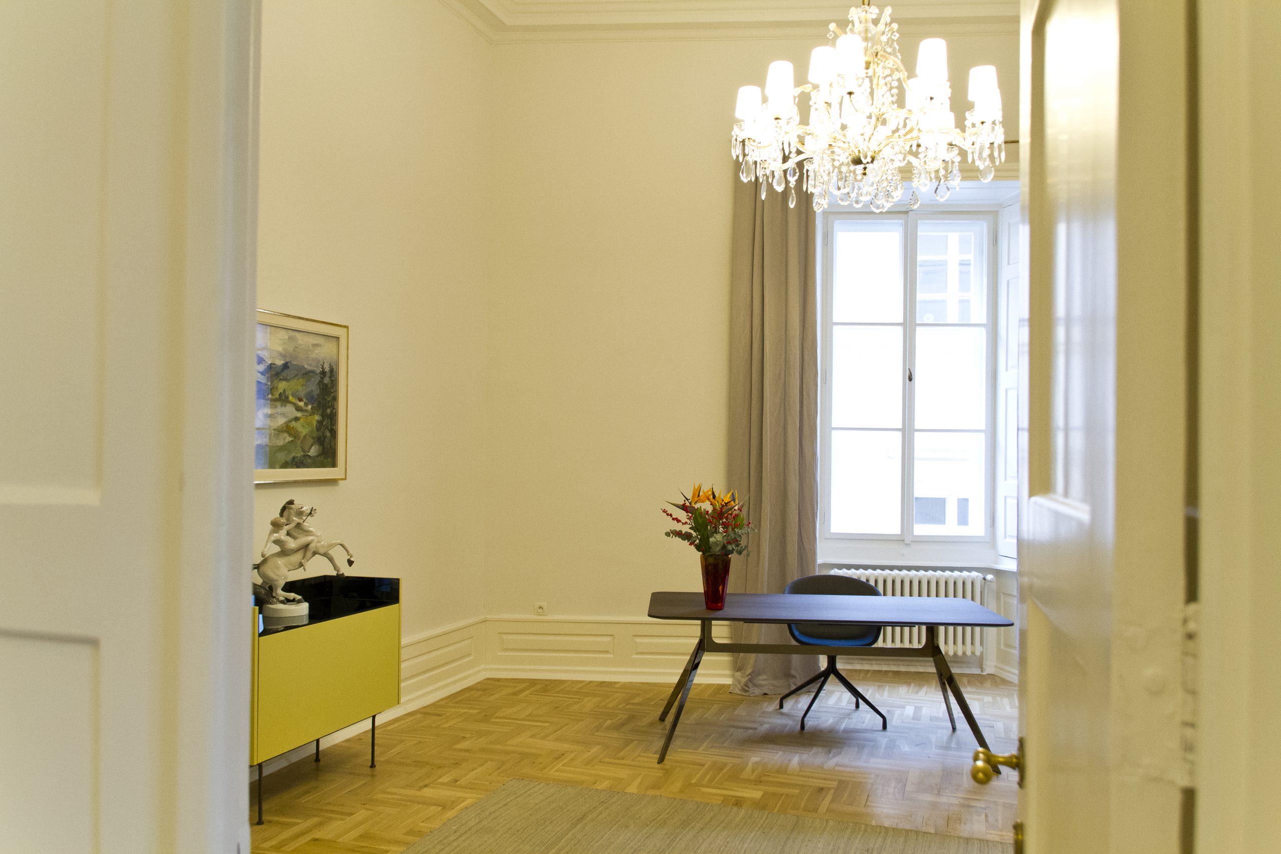 Ein Zimmer in der Bayerischen Repräsentanz in Prag: Ein kleiner Schrank links. Über den kleinen Schrank hängt ein Bild an der Wand. Rechts steht vor dem Fenster ein Tisch.