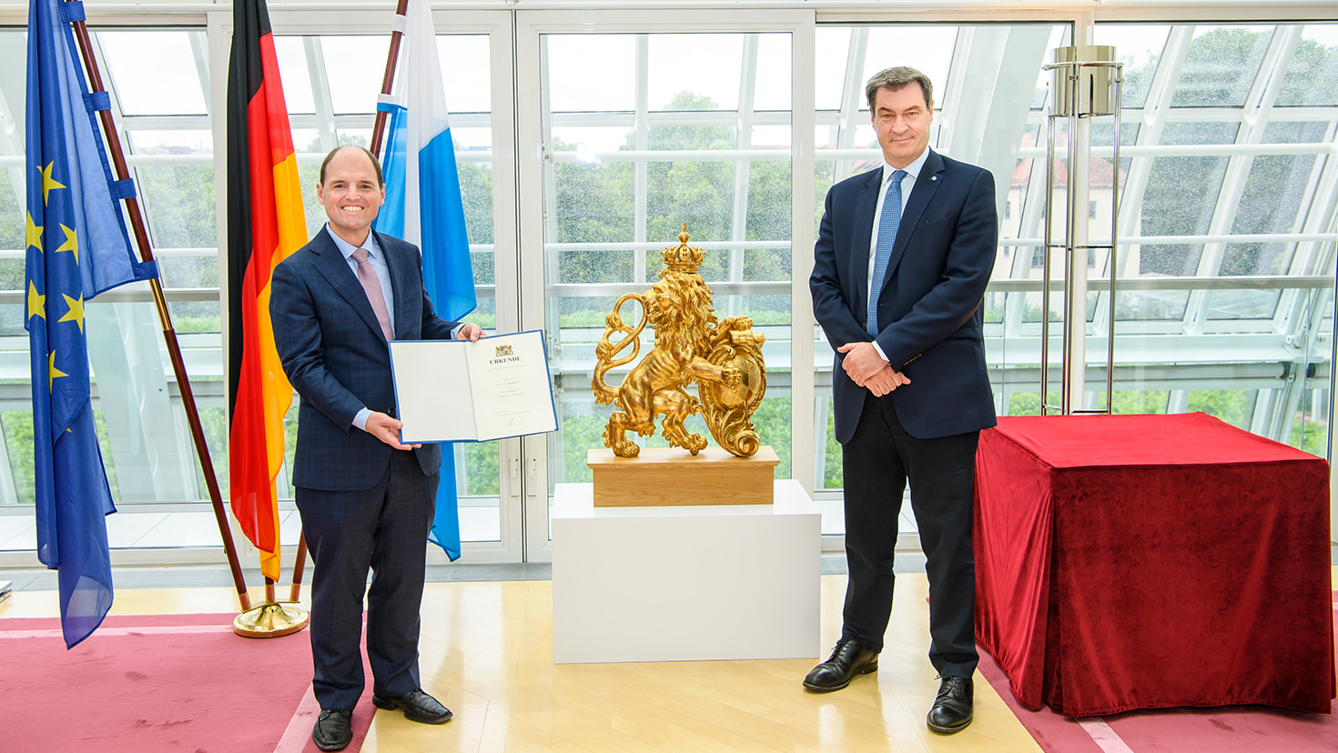 Der Bürgerbeauftragte der Bayerischen Staatsregierung, Michael Hofmann, MdL (links), und Ministerpräsident Dr. Markus Söder, MdL (rechts), nach der Urkundenübergabe in der Staatskanzlei. Im Hintergrund sind links die Fahnen der Europäischen Union, der Bundesrepublik Deutschland und des Freistaats Bayern sowie in der Mitte eine goldene Statue des bayerischen Löwen zu sehen.