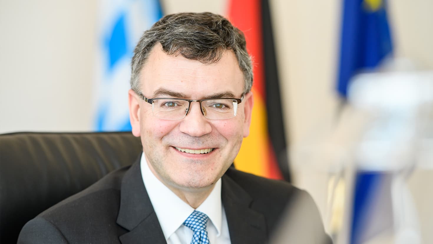 Staatsminister Dr. Florian Herrmann, MdL, sitzt an seinem Schreibtisch in der Staatskanzlei. Im Hintergrund sind die Flaggen des Freistaats Bayern, der Bundesrepublik Deutschland und der Europäischen Union (von links nach rechts) zu sehen.