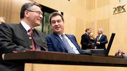 Staatsminister Dr. Florian Herrmann (links) und Ministerpräsident Dr. Markus Söder (rechts) sitzen nebeneinander und unterhalten sich lächelnd miteinander im Sitzungssaal des Bundesrats in Berlin.