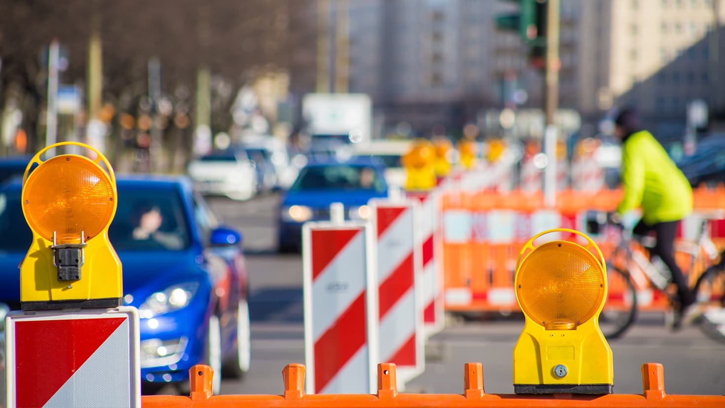 Fahrzeuge fahren an einer Straßenbaustelle in der Stadt vorbei. Ein Radfahrer (rechts) will die Straße überqueren. © Rainer Fuhrmann - stock.adobe.com