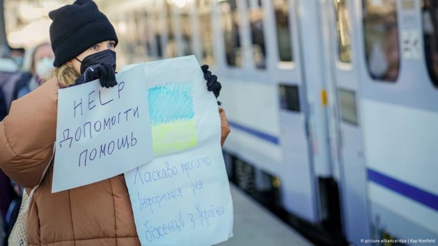 Eine Frau steht am Bahnhof. Sie hält ein Plakat mit Hilfsangebot und ukrainischer Flagge hoch.