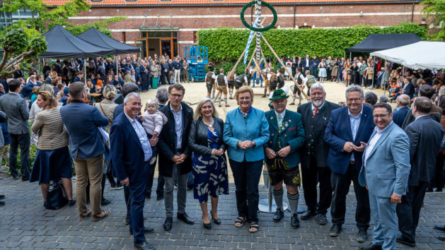 Die Bayerische Europaministerin steht gemeinsam mit Ehrengästen auf den Treppenstufen der Vertretung. Im Hintergrund sieht man die Trachtler beim Maibaumaufstellen.