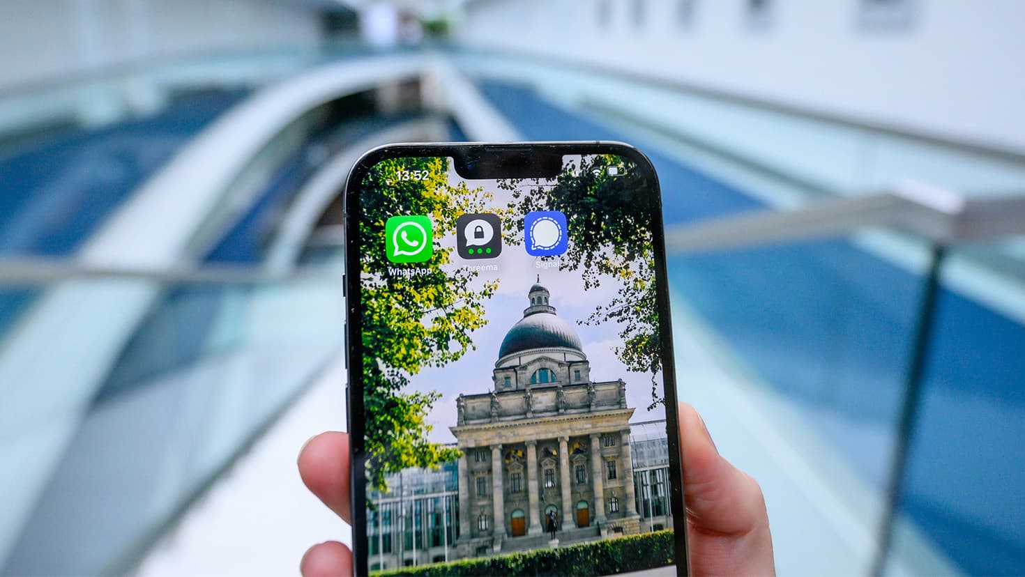 Logos der Apps WhatsApp, Signal und Threema auf einem Handy mit Desktopbild der Staatskanzlei.