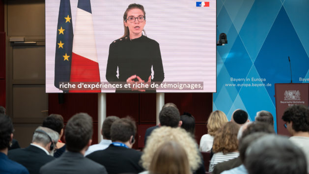 Viele Menschen im Saal schauen auf einen Bildschirm, wo eine Frau neben der EU- und der französischen Flagge zu sehen ist.