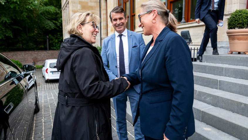 Die beiden Damen schütteln sich lächelnd die Hand. Minister Beißwenger ist in der Mitte zu sehen und lächelt. Rechts geht es die Treppe hinauf.